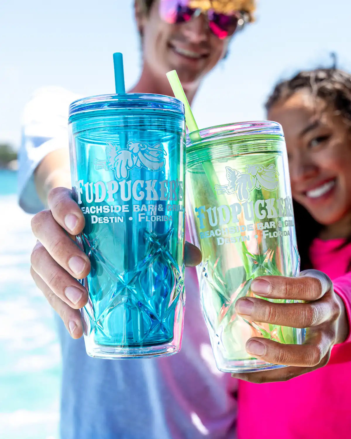 Two people holding blue and green tumblers with 'Fudpucker's' branding, outdoors.